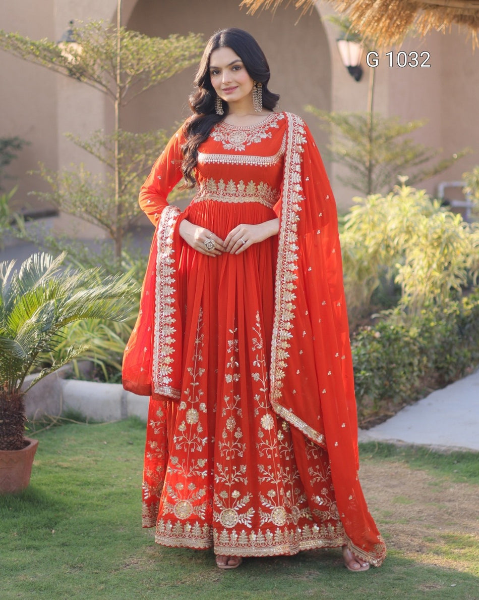 A woman models a traditional red Indian ethnic outfit with intricate gold embroidery, featuring a long kurta, matching dupatta, and a red skirt, set against an outdoor garden background.