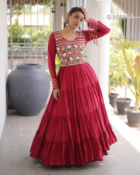 A woman models a long-sleeved red maxi dress with a heavily embroidered bodice and tiered skirt, standing on a covered walkway.