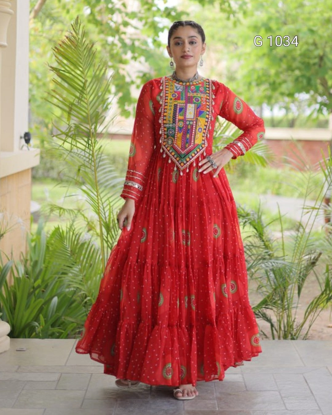 A woman modeling a red long Anarkali gown with a heavily embroidered multicolor yoke and scattered gold motifs, standing outdoors on a tiled walkway with green plants in the background.