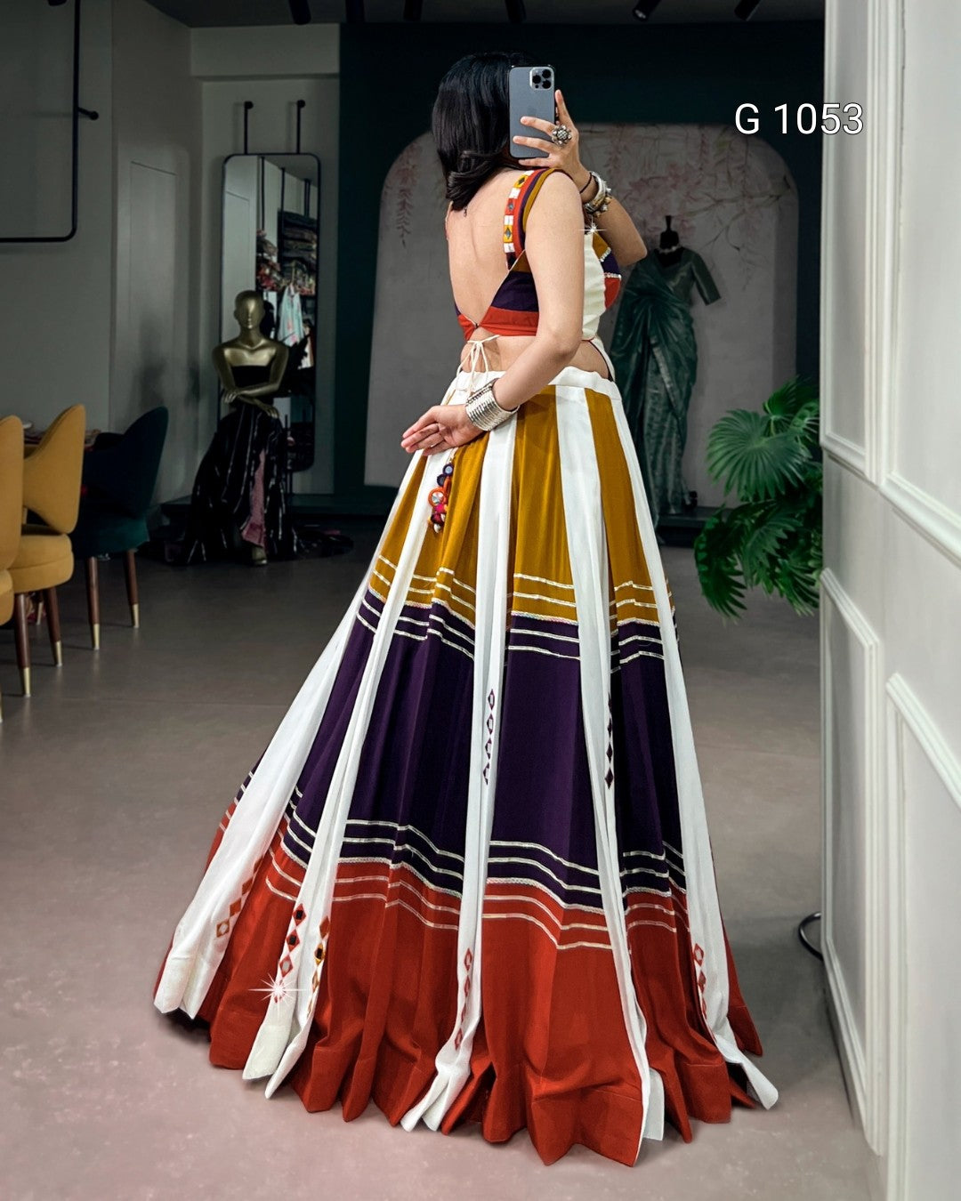 A woman takes a mirror selfie in a boutique wearing a traditional Indian lehenga choli. The lehenga features vertical stripes of white, mustard yellow, deep purple, and rust orange with intricate embroidery.
