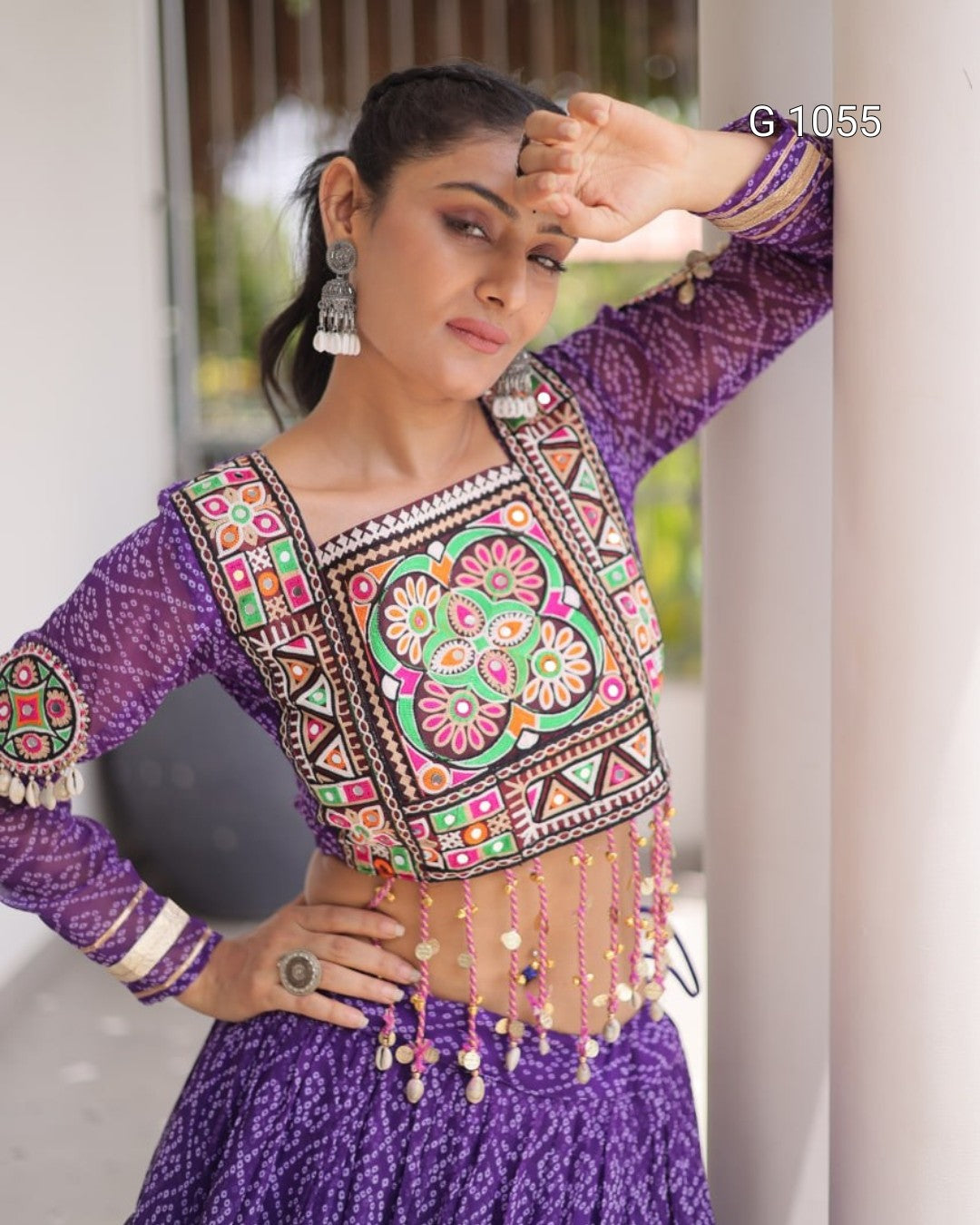 A woman wearing a purple patterned traditional outfit with a heavily embroidered multicolor crop top and matching skirt, posing outdoors against a light-colored wall.
