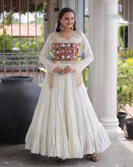 A woman models a traditional long white anarkali-style dress with a heavily embroidered multicolor bodice panel featuring pink, orange, and gold patterns, set against a porch background with white pillars.