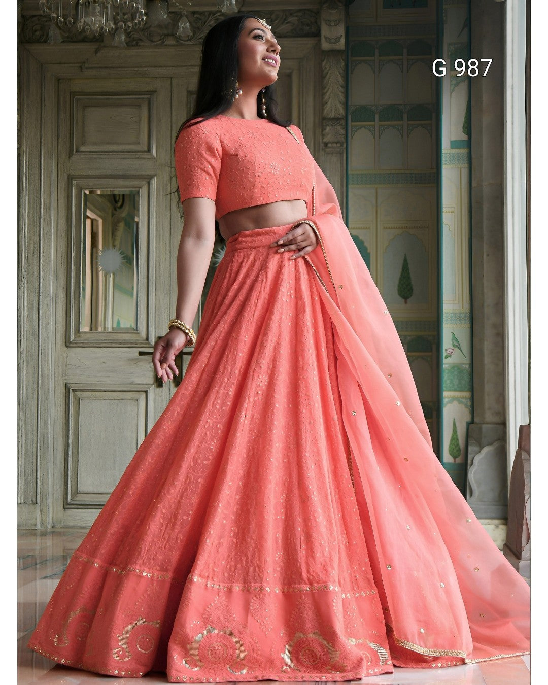 A woman models a peach-colored traditional Indian ethnic wear set consisting of a short-sleeve crop top, flared skirt, and dupatta, set against an ornate architectural background.