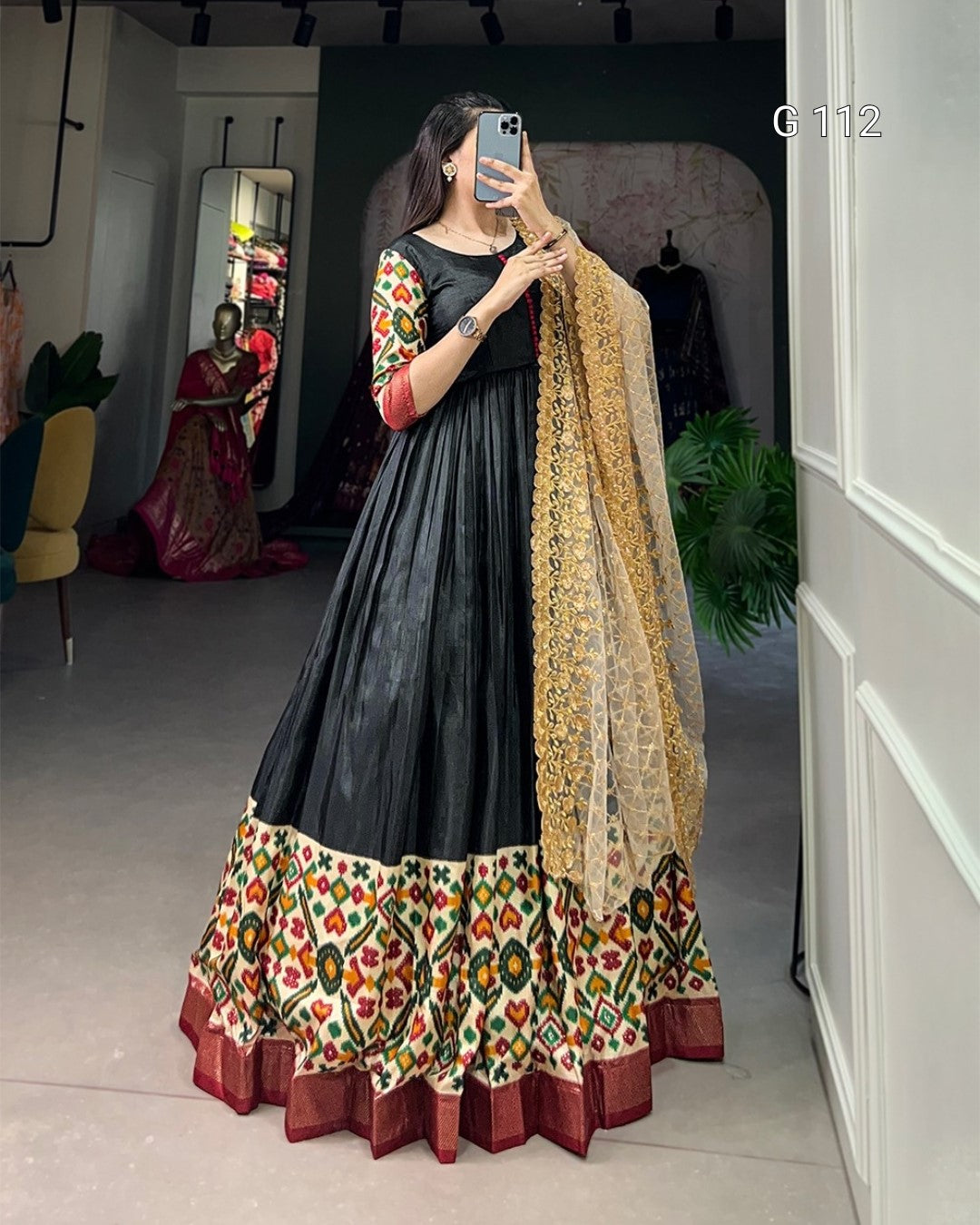 Woman taking a mirror selfie wearing a black dress with colorful patterns and a yellow dupatta.