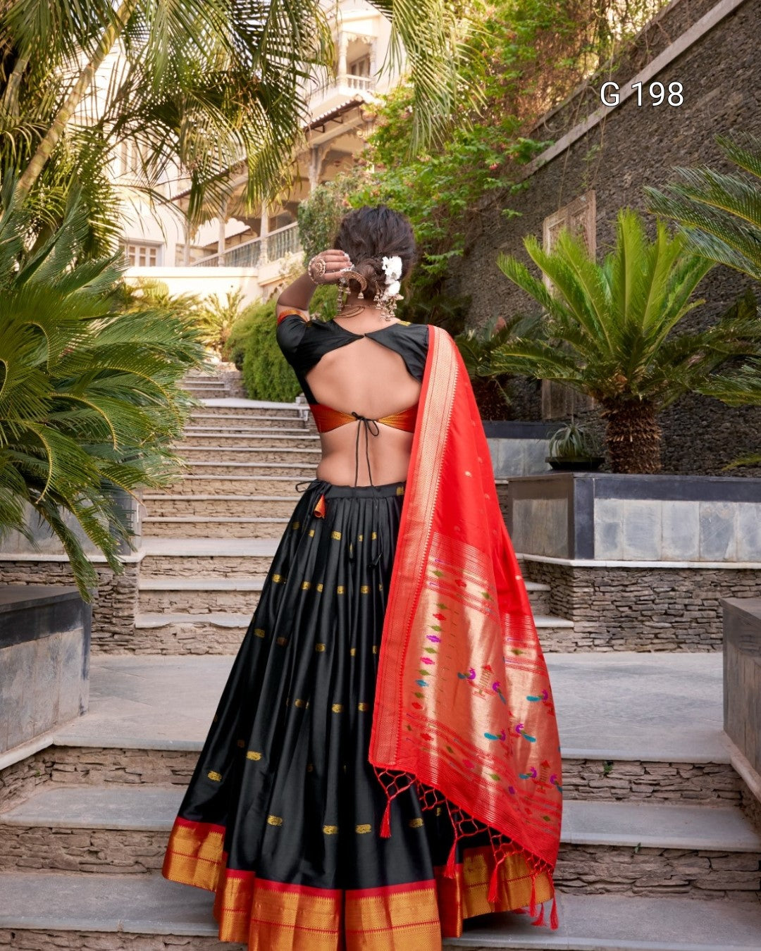Woman in a black saree with a red blouse standing outdoors with plants and steps in the background