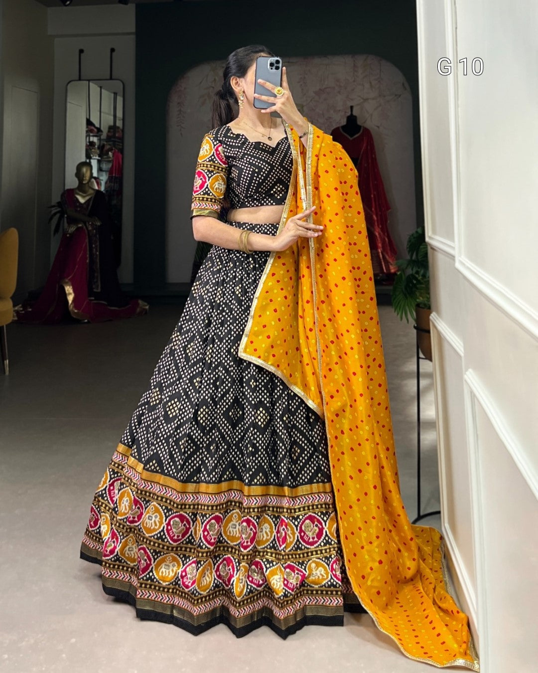 Woman in a traditional outfit with a black and colorful patterned dress and yellow dupatta, standing in a room.
