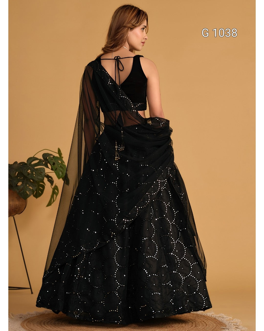 A woman modeling a black embellished lehenga choli dress from the back, standing against a warm beige studio backdrop with a small plant at the side. The text 'G 1038' appears in the top right.