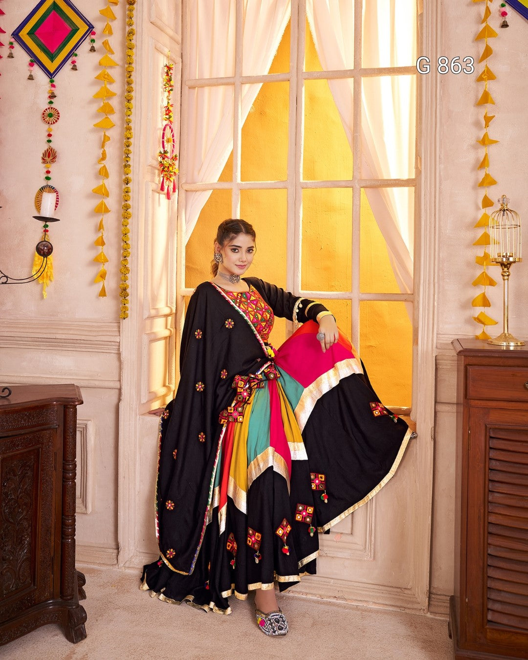 Woman in a colorful saree sitting in a decorated room with yellow curtains and festive decorations.