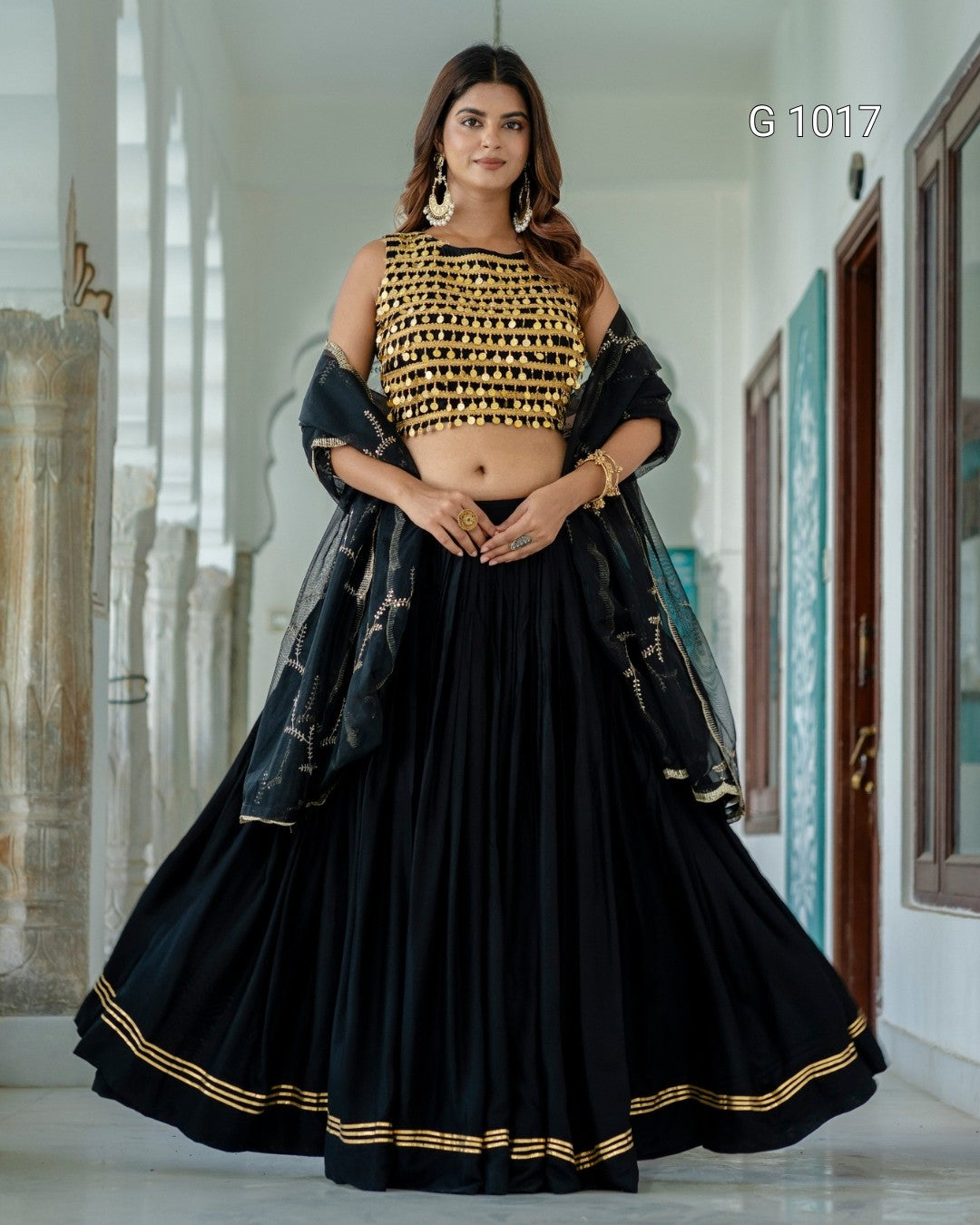 A woman models a black and gold traditional Indian outfit consisting of a heavily embroidered black crop top, a full black flared skirt with gold border detailing, and a sheer black dupatta draped over her shoulders, set against a bright hallway background.