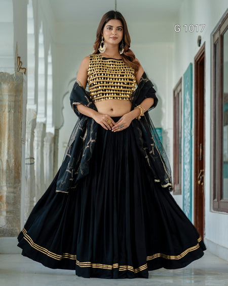 A woman models a black and gold traditional Indian outfit consisting of a heavily embroidered black crop top, a full black flared skirt with gold border detailing, and a sheer black dupatta draped over her shoulders, set against a bright hallway background.