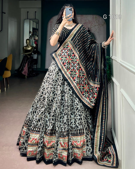 A woman takes a mirror selfie wearing an ornate black and gray printed lehenga choli set with a heavily embroidered multicolor dupatta. The set features intricate red, green, and gold patterns on a black base, with a black and gray floral print on the skirt.