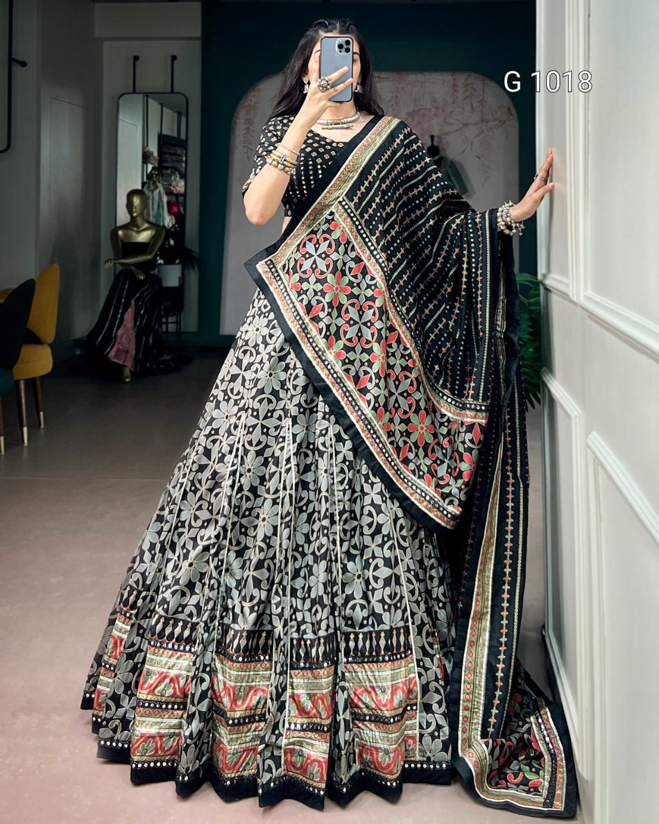 A woman takes a mirror selfie wearing an ornate black and gray printed lehenga choli set with a heavily embroidered multicolor dupatta. The set features intricate red, green, and gold patterns on a black base, with a black and gray floral print on the skirt.