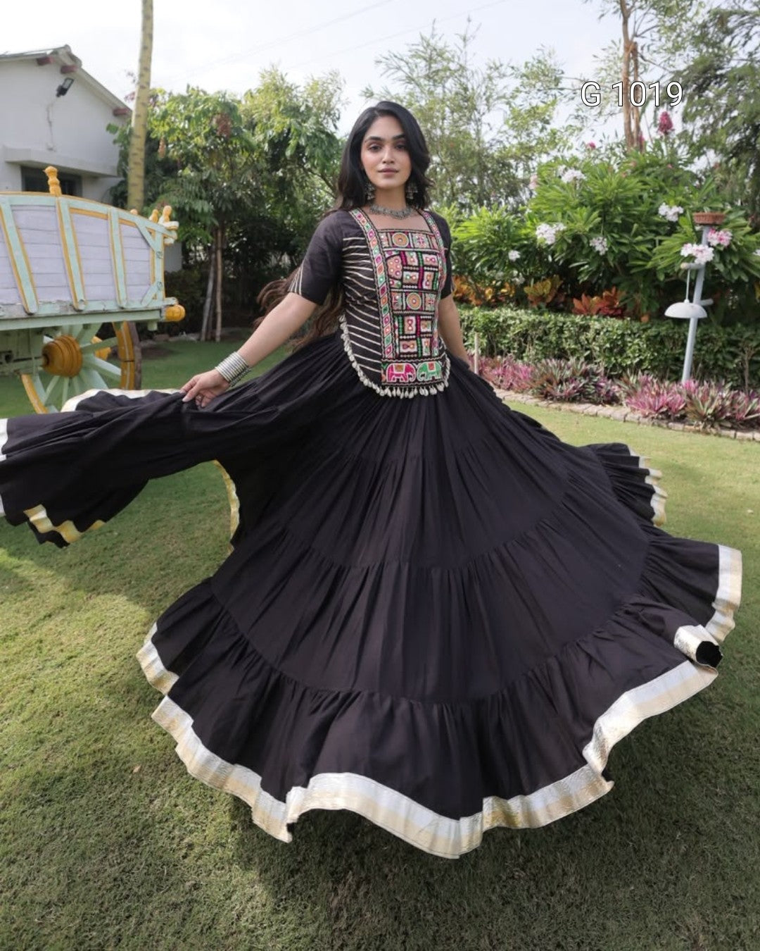 A woman outdoors on a lawn wearing a black traditional dress with a heavily embroidered neckline and a very full flared skirt with cream trim, holding the skirt out to show its volume.