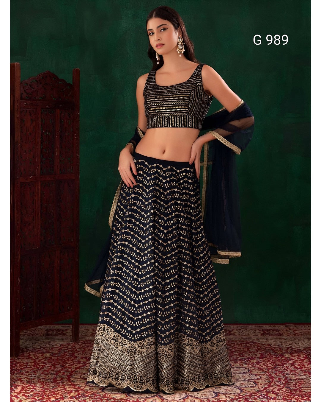 A woman models a black and gold embellished lehenga choli set against a dark green backdrop. The outfit features a black sleeveless crop top with gold embroidery, a matching flared skirt with intricate gold patterns, and a sheer black dupatta with gold borders.