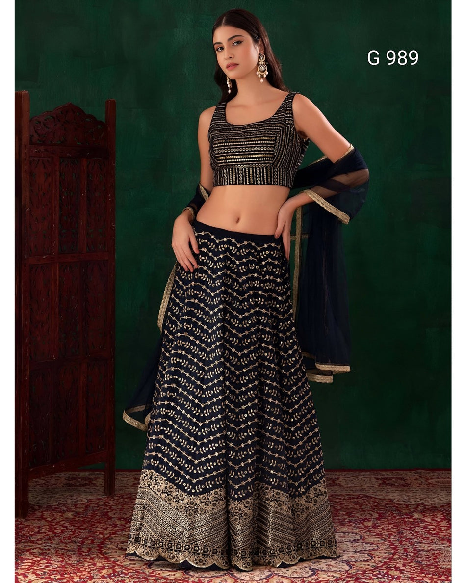 A woman models a black and gold embellished lehenga choli set against a dark green backdrop. The outfit features a black sleeveless crop top with gold embroidery, a matching flared skirt with intricate gold patterns, and a sheer black dupatta with gold borders.