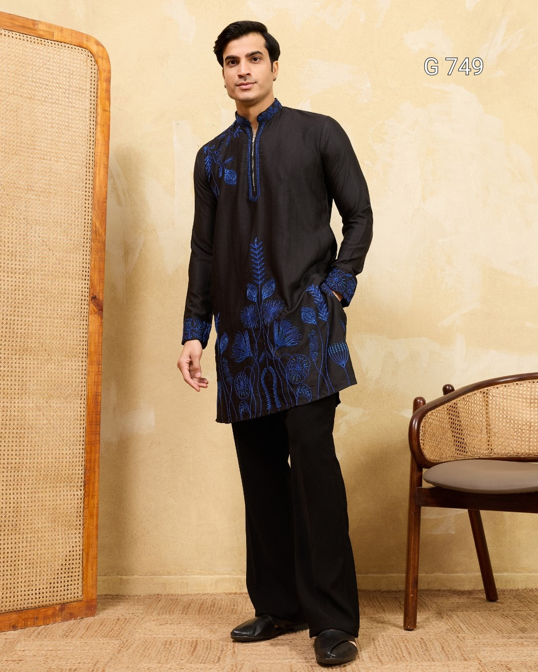 Man wearing a black kurta with blue embroidery standing against a beige wall.