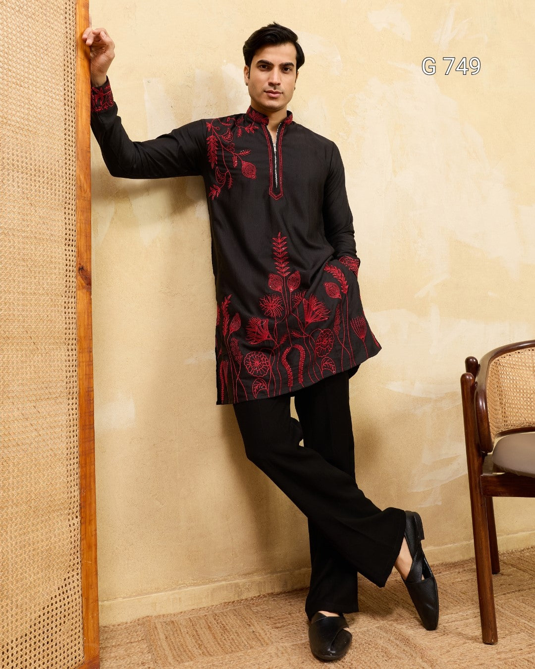 Man wearing a black kurta with red patterns against a beige wall.