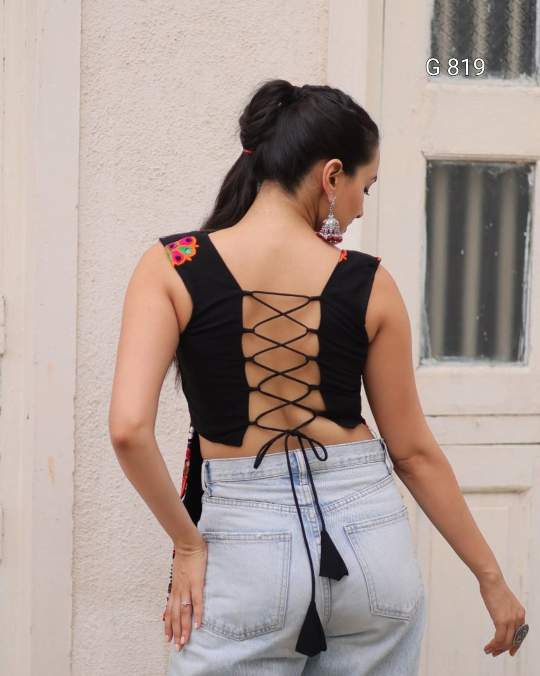 Woman wearing a black lace-up top and light blue jeans against a beige wall.