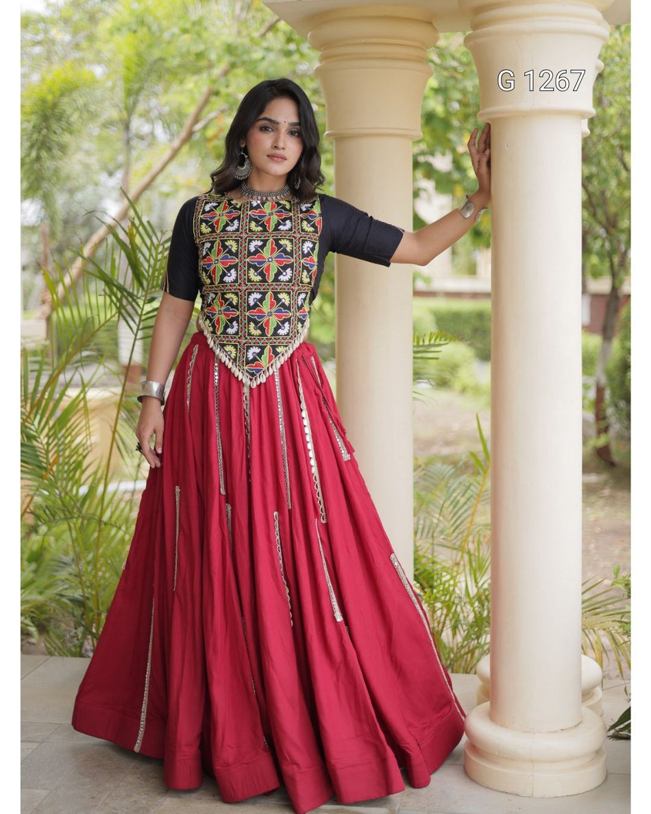 A woman models a traditional Indian Anarkali gown with a black short-sleeve top and a voluminous red skirt featuring gold and silver mirror work and embroidery. She is posing outdoors between two cream-colored pillars with green foliage in the background.
