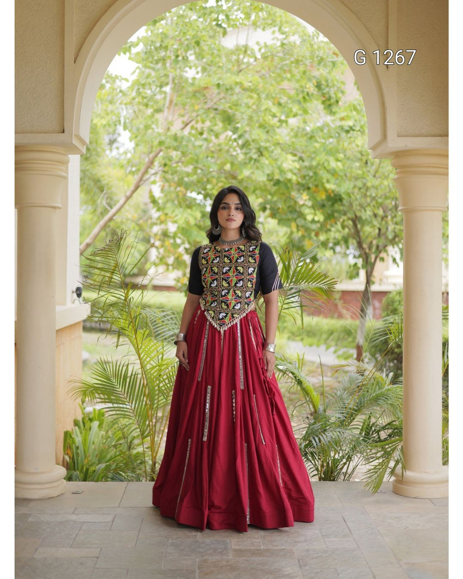 A woman stands in a stone archway wearing a long red anarkali-style gown with a black short-sleeve top and a heavily embroidered gold, black, and red bodice. Green garden foliage is visible behind her.