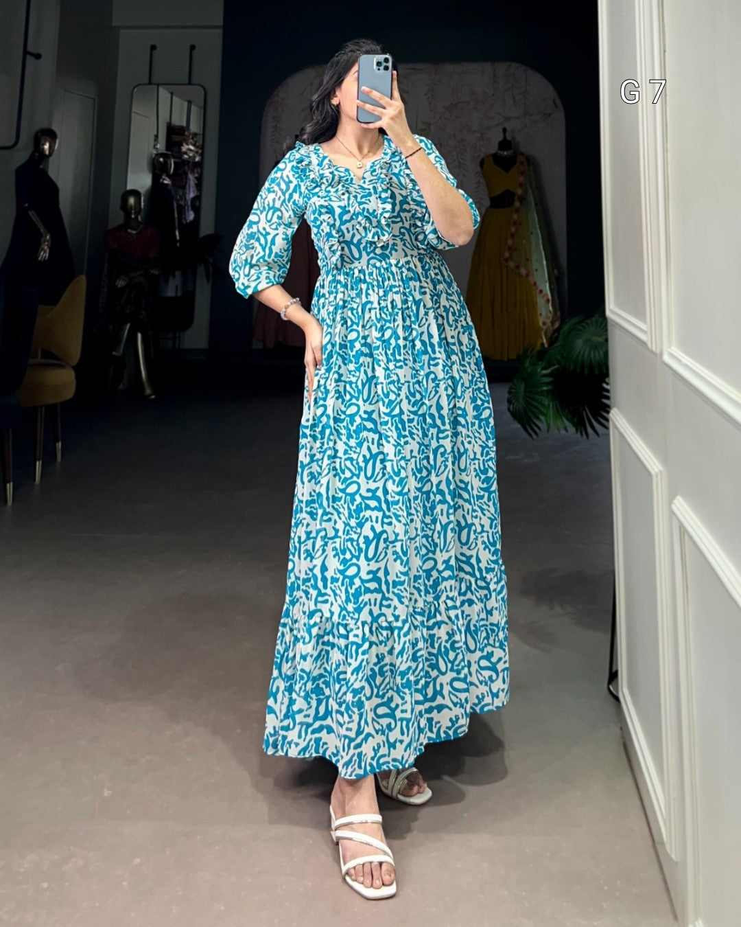 Woman in a blue floral dress taking a mirror selfie in a room with gray walls and a white door.