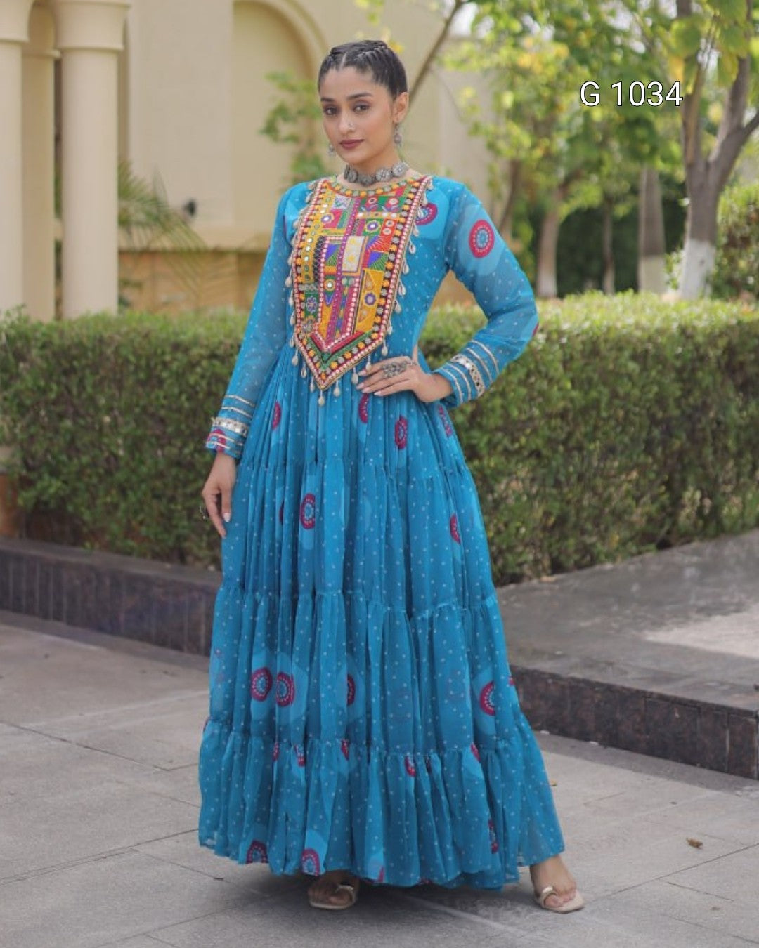 A woman modeling a traditional blue Anarkali gown with a heavily embroidered multicolor yoke and printed motifs on the tiered skirt, standing outdoors in front of a building and hedges.