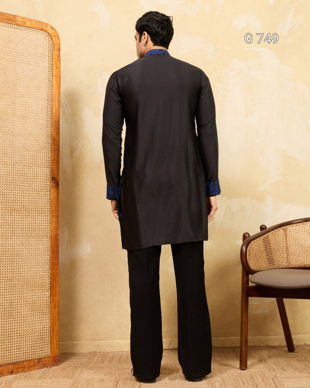 Person wearing a black kurta with blue accents standing against a beige wall.