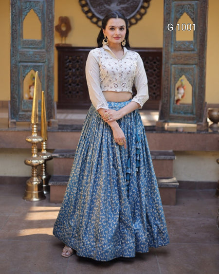 A woman models a traditional Indian ethnic outfit consisting of a white embroidered crop top and a long blue floral printed skirt, standing in front of an ornate architectural background with brass lamps.