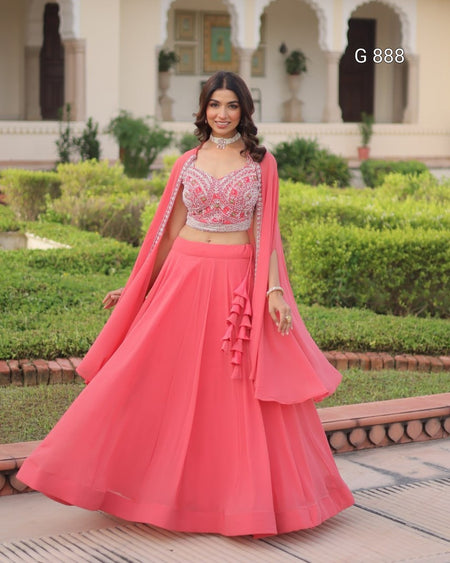 Woman in a pink traditional outfit standing outdoors with greenery and architecture in the background.