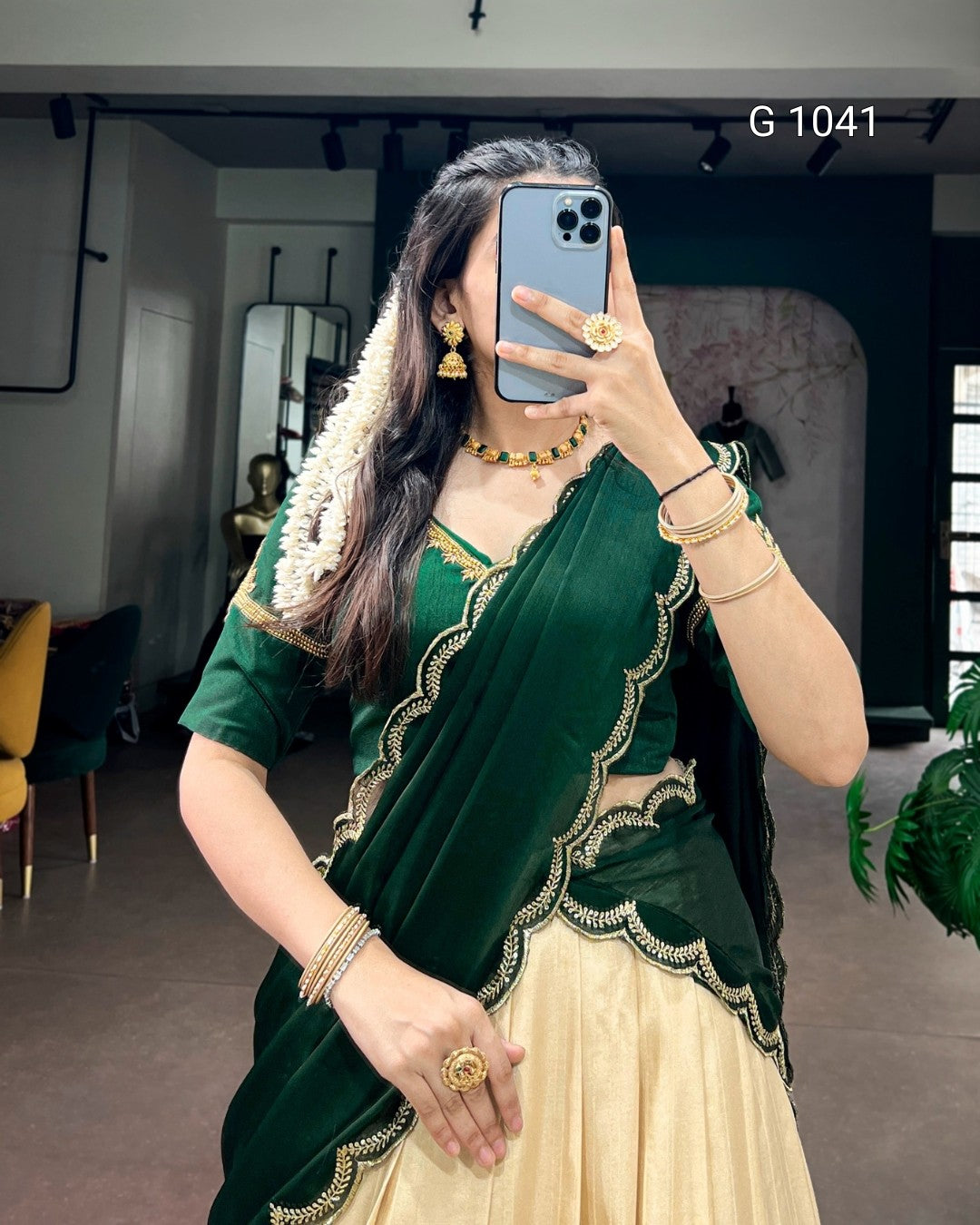 A woman takes a mirror selfie wearing a traditional Indian outfit consisting of a dark green velvet sari with gold scalloped borders, a matching green blouse with gold embroidery, and a cream-colored flared skirt. She is accessorized with gold jewelry, including earrings, a necklace, rings, and bangles.