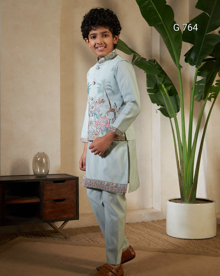 Young boy in traditional embroidered outfit standing in a room with a plant and wooden cabinet.