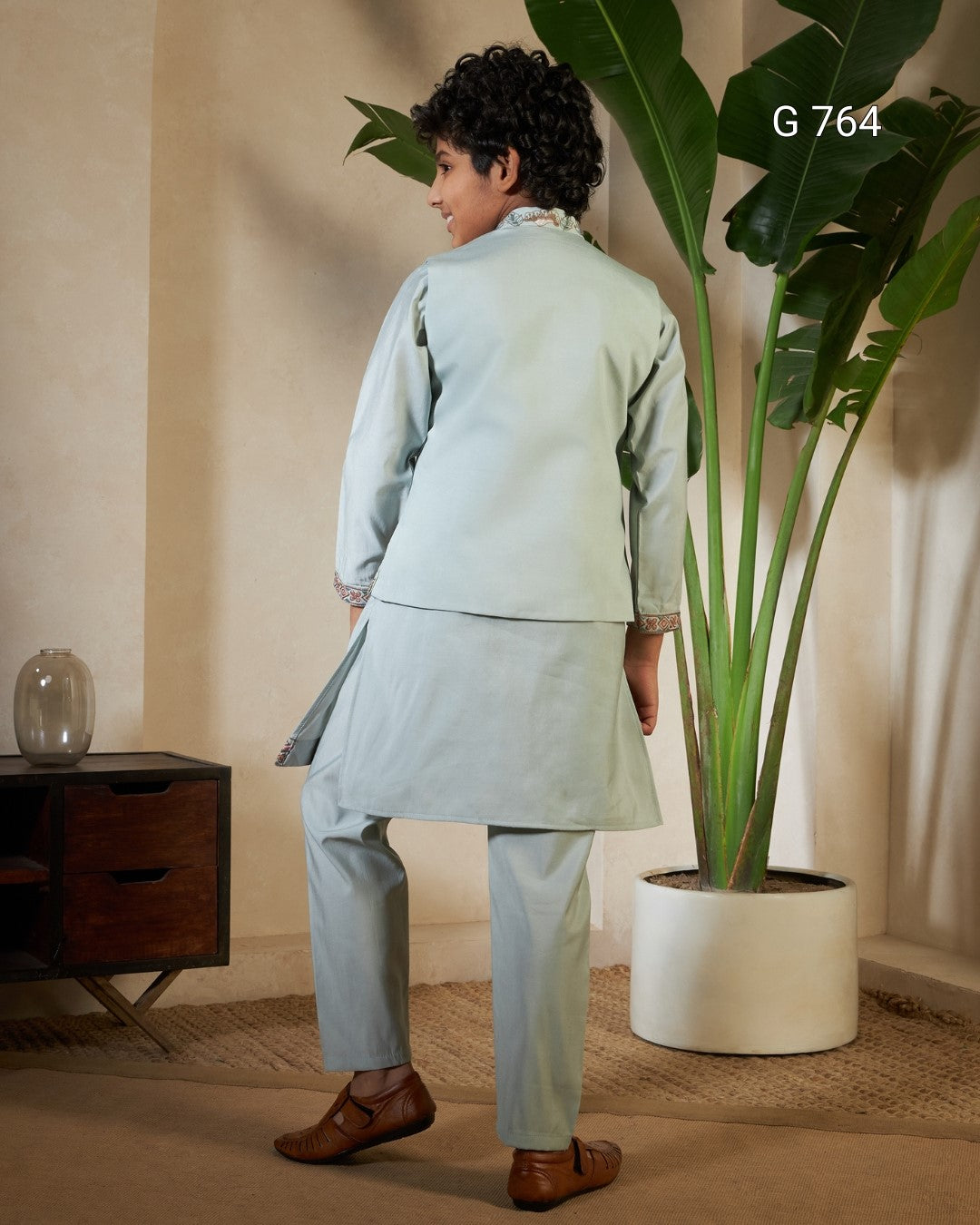 Person wearing a light blue traditional outfit standing in a room with a plant and wooden cabinet.