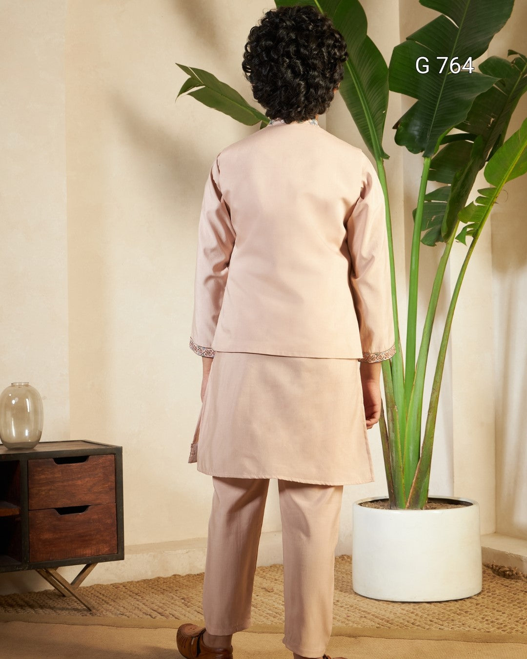 Person wearing a beige outfit standing next to a large plant indoors.
