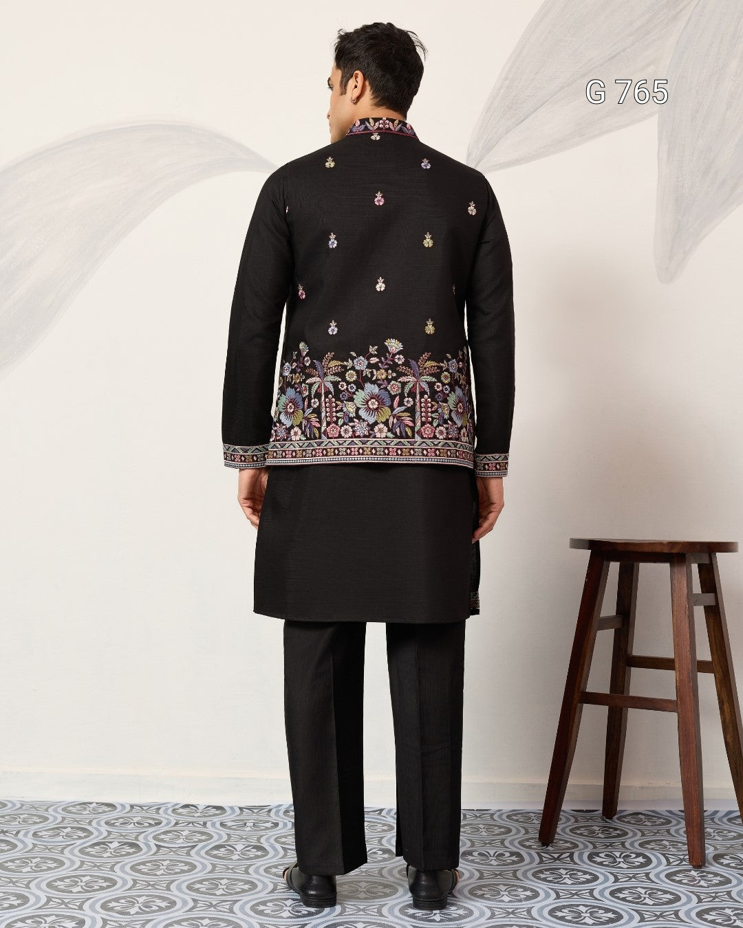 Man wearing a black embroidered traditional outfit standing in a room with a white wall and wooden stool.