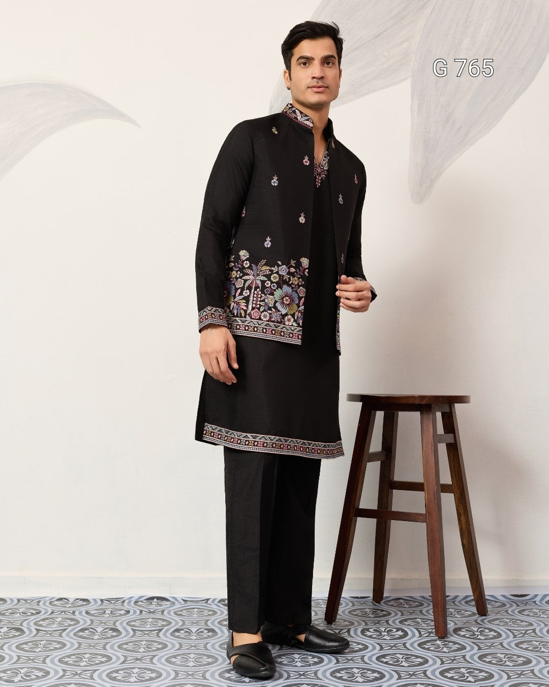 Man wearing a black traditional outfit with intricate patterns in a minimalistic setting.