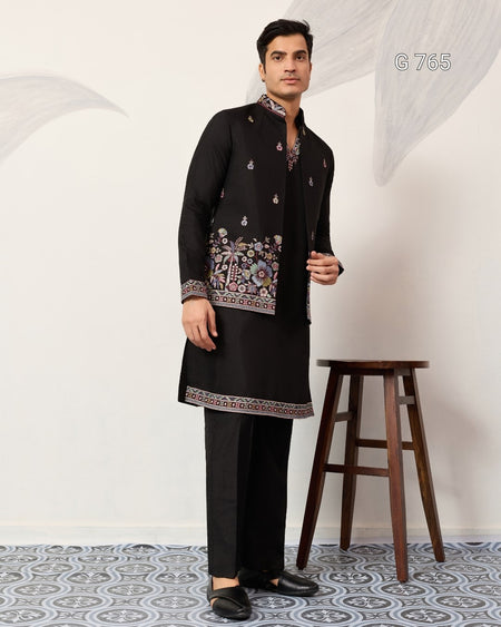 Man wearing a black traditional outfit with intricate patterns in a minimalistic setting.