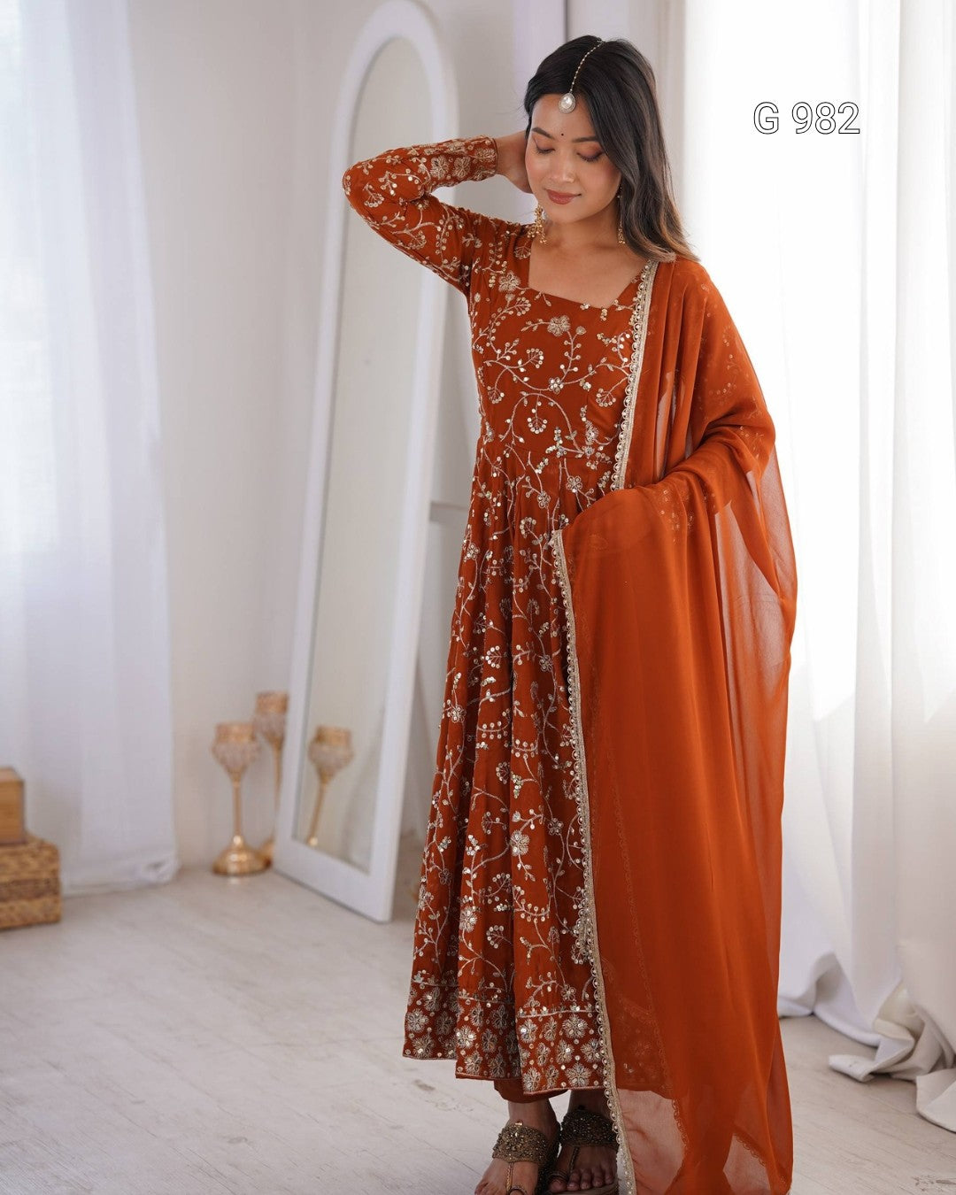 A woman models a rust-orange long-sleeve Anarkali gown with intricate white floral embroidery and a matching sheer dupatta, standing in a bright, minimalist room with white curtains and a mirror.