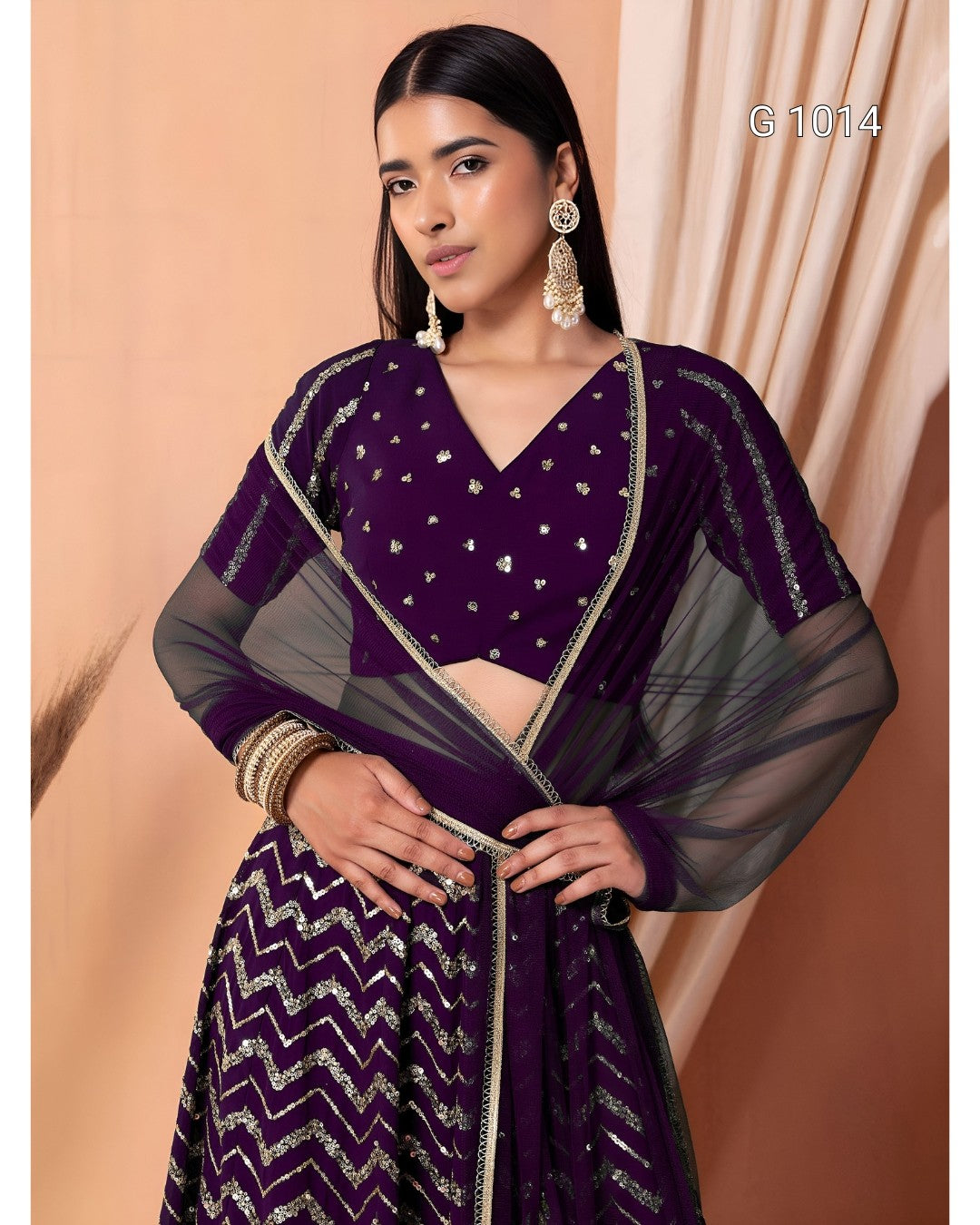 A woman modeling a deep purple traditional outfit with silver embellishments, sheer sleeves, and a chevron-patterned skirt, posed against a peach-colored draped backdrop.