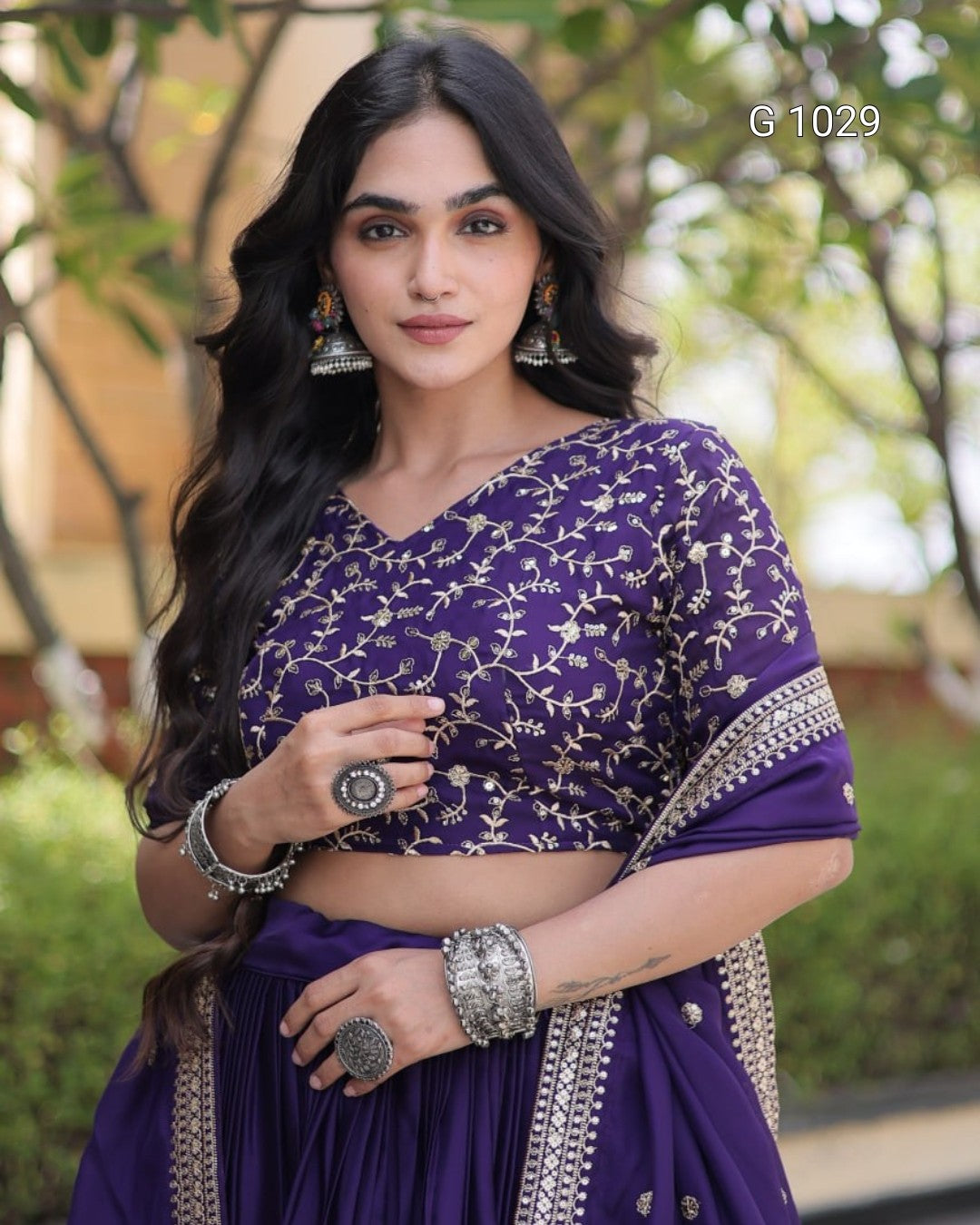A woman wearing a purple traditional outfit with silver floral embroidery on the blouse, paired with a matching pleated skirt and draped dupatta, posed outdoors with a blurred green background.