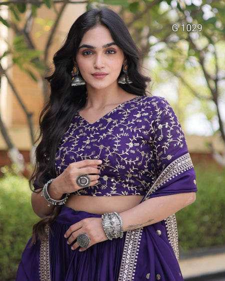 A woman wearing a purple traditional outfit with silver floral embroidery on the blouse, paired with a matching pleated skirt and draped dupatta, posed outdoors with a blurred green background.