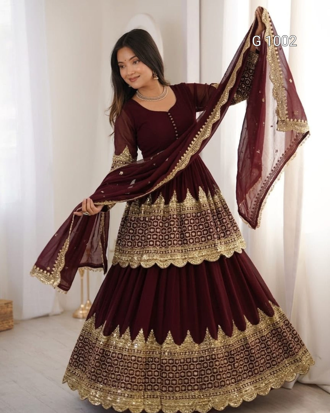 A woman modeling a traditional maroon and gold georgette lehenga choli set with a matching dupatta, standing in a bright indoor setting.