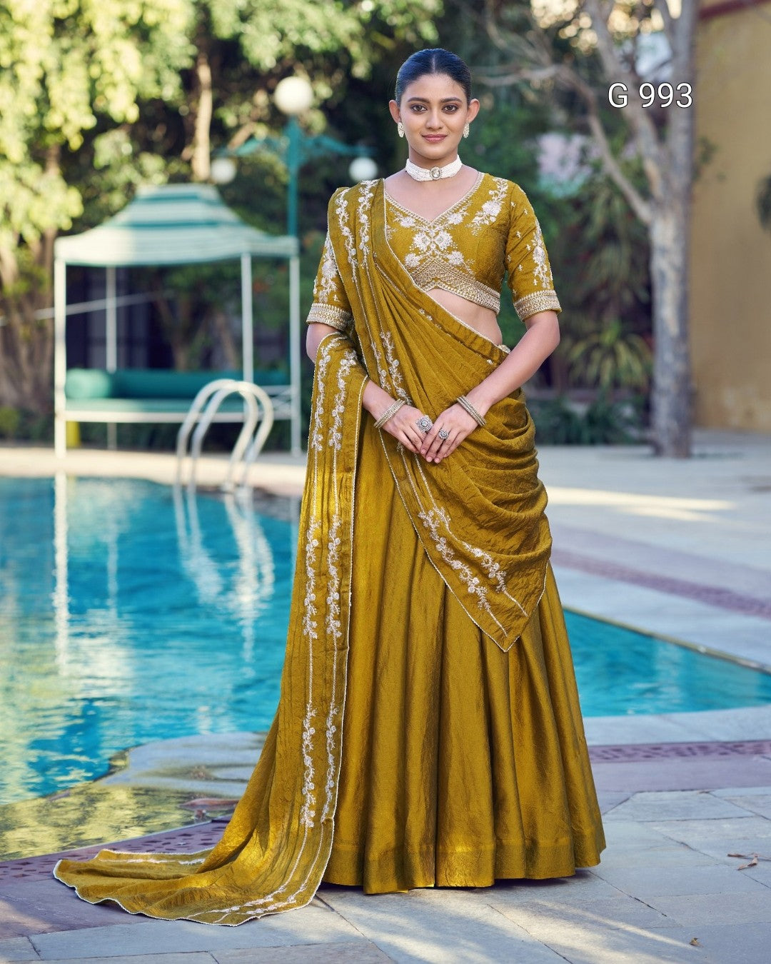 A woman models a mustard yellow ethnic lehenga choli set with white embroidery, standing by a swimming pool in an outdoor setting.