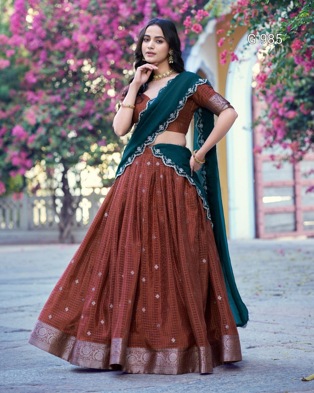 A woman modeling a traditional Indian lehenga choli set outdoors. The outfit features a rust brown flared skirt with silver embroidery, a matching brown blouse, and a dark teal green dupatta with silver border detailing.