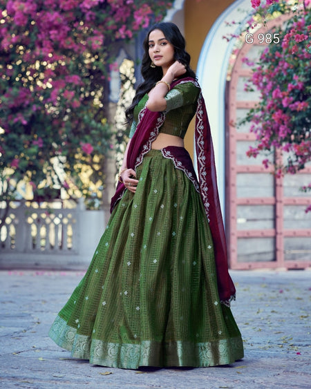 A woman wearing an olive green embroidered lehenga choli set with a deep maroon dupatta, standing outdoors in front of a stone archway with blooming pink flowers.