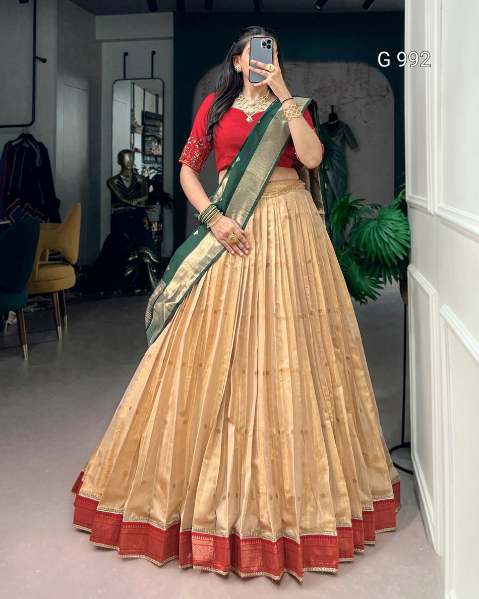 A woman takes a mirror selfie wearing a traditional Indian outfit consisting of a beige pleated skirt with red border, a red blouse, and a green and gold sash.