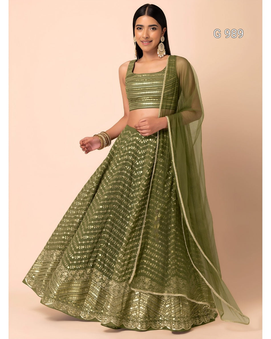 A woman modeling an olive green embroidered lehenga choli set with a matching sheer dupatta, standing against a solid pale peach background.