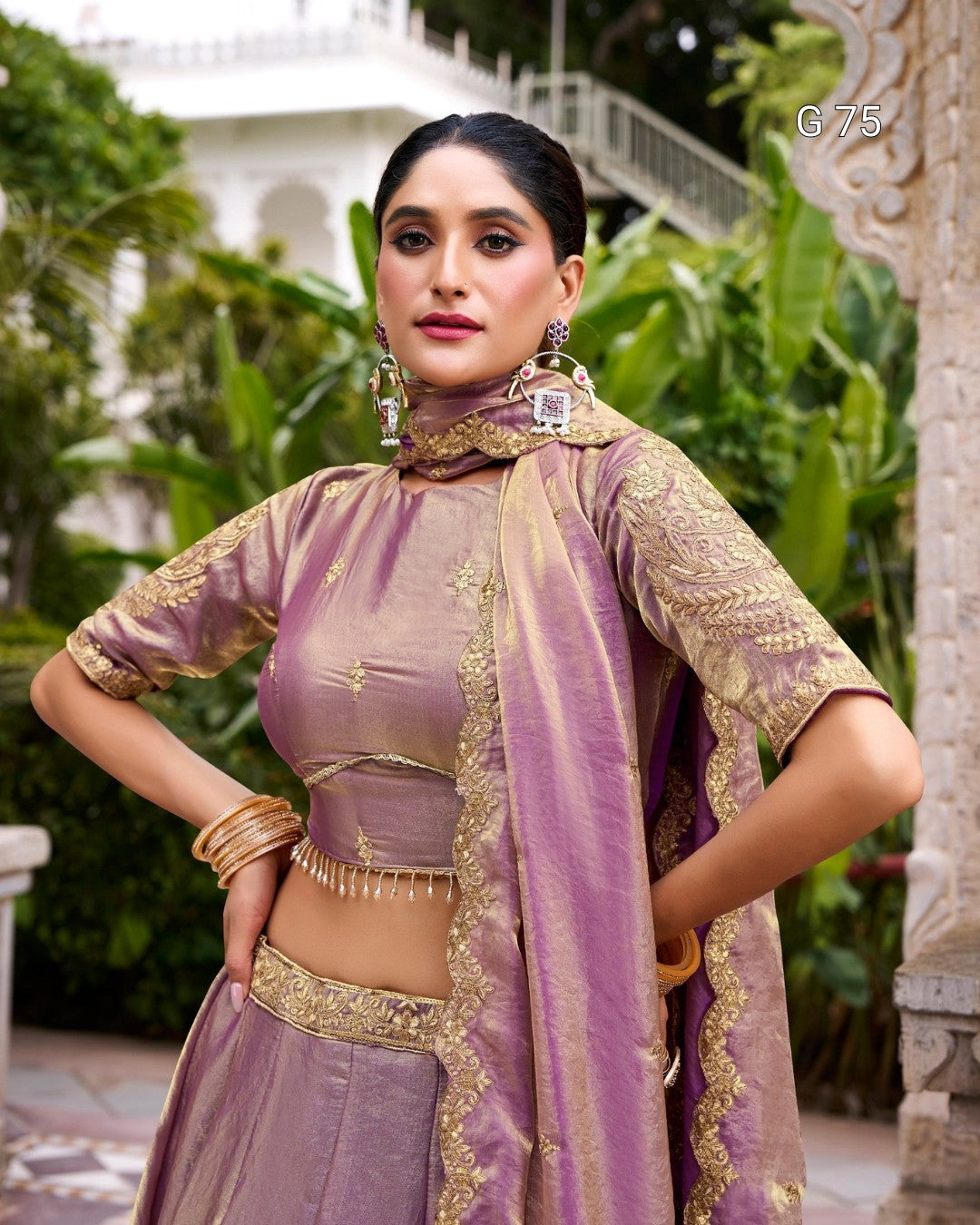 A woman modeling a lavender purple traditional Indian outfit with gold embroidery, featuring a high-necked crop top, matching skirt, and a coordinating dupatta, posed outdoors with a white building and green foliage in the background.
