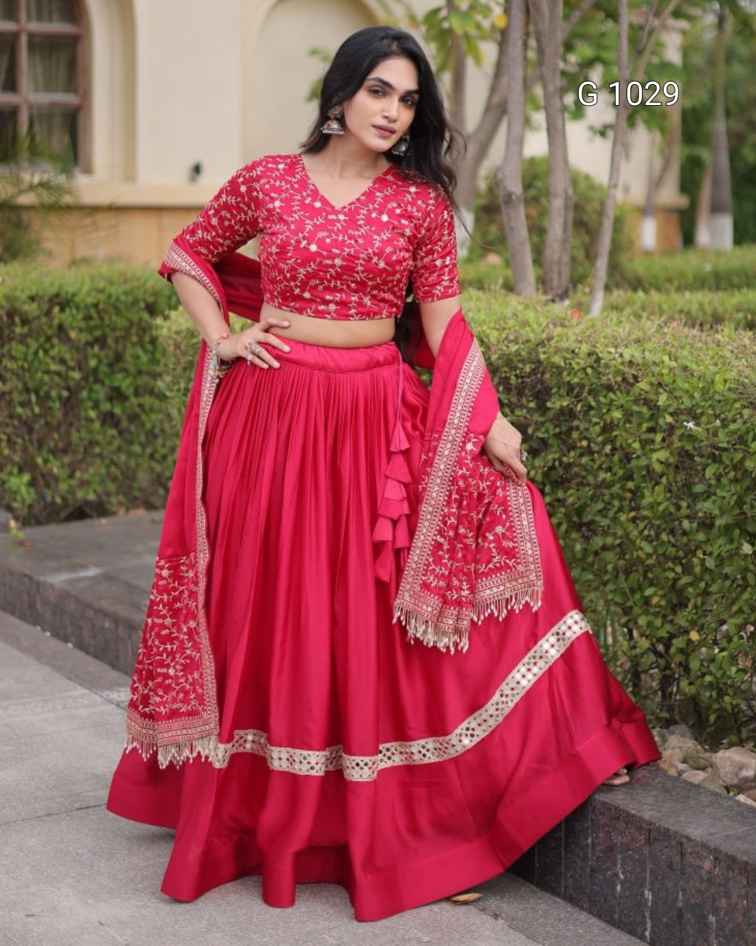 A woman models a bright red lehenga choli with intricate gold embroidery, standing outdoors on a stone pathway with green hedges in the background.