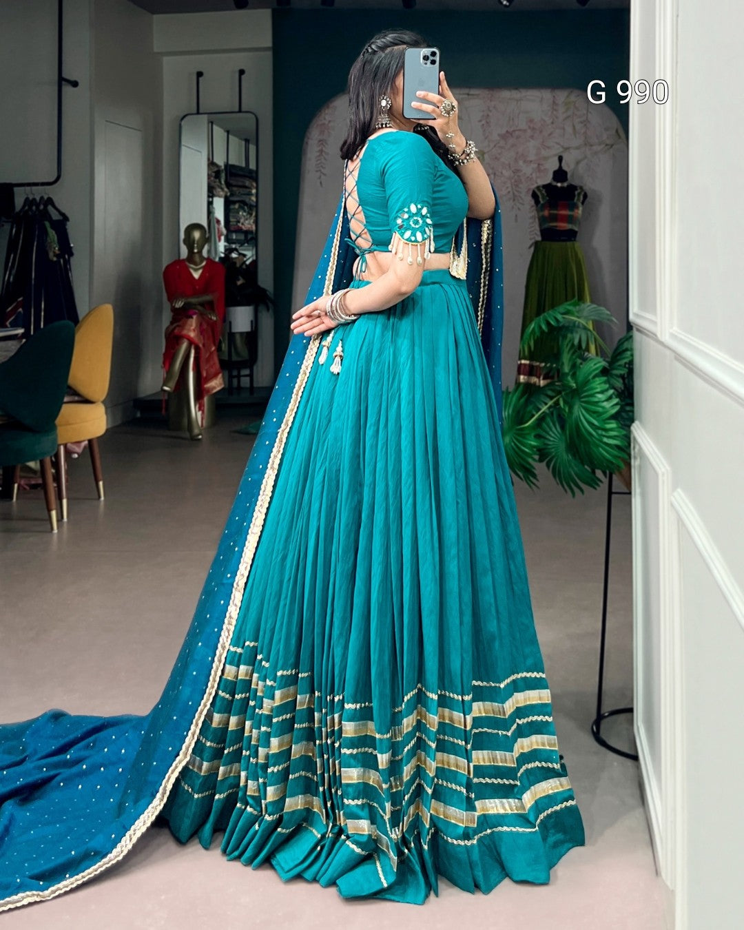 A woman wearing a teal blue traditional Indian lehenga choli dress with gold zari embroidery, taking a mirror selfie in a boutique showroom.