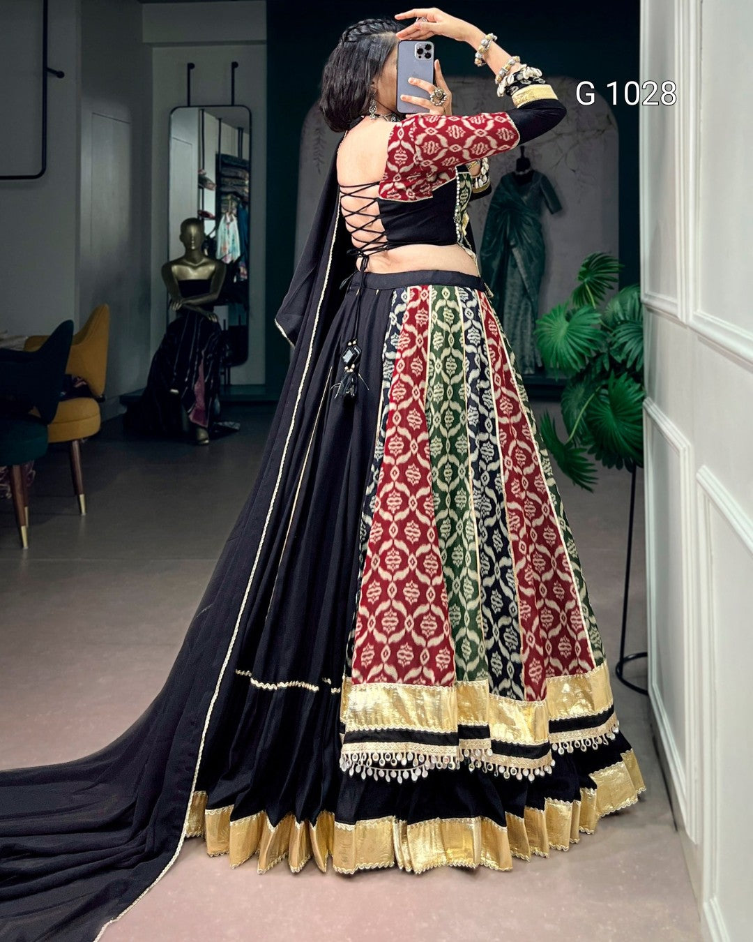 A woman takes a mirror selfie in a boutique wearing a traditional Indian lehenga choli outfit. The outfit features a black base with intricate red, green, and gold patterns on the skirt, a red and gold patterned blouse, and a long black dupatta with gold borders.